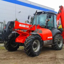 Czerwona koparko-ładowarka Manitou na placu budowy, widok z boku, z innymi maszynami w tle. Widoczne detale opon i logo firmy.