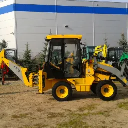 Żółta koparko-ładowarka z numerem 1374 na piaszczystym podłożu, na tle budynku z niebieskimi pasami i rzędów małych drzewek.