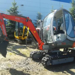 Czerwono-szara minikoparka Neuson 2203 na gąsienicach, z kabiną, na placu budowy. W tle inne maszyny i drzewa iglaste. Jasne oświetlenie.