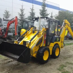 Żółta miniładowarka JCB z czarną łyżką na tle innych maszyn budowlanych i budynku. Widoczne drzewa iglaste i liściaste. Lekko pochmurna pogoda.