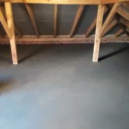 Szara, gładka powierzchnia w pomieszczeniu ze skośnym dachem, widoczne drewniane belki konstrukcyjne i słupy podtrzymujące dach.