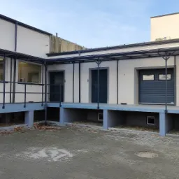 Rampa załadunkowa z trzema wejściami: dwoje drzwi i segmentowa brama garażowa w kolorze grafitowym, zadaszona konstrukcja metalowa, ściany w kolorze kremowym.