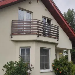 Dom jednorodzinny z beżową elewacją i balkonem z brązową, poziomą balustradą, widok z ogrodu z kwitnącymi różami.