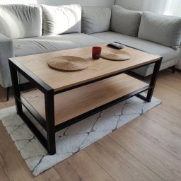 FilART - Construction Damian Filar - Stylowy stolik kawowy z drewnianym blatem i czarną metalową ramą, na puszystym dywanie w salonie, obok szarej sofy. Widoczne dekoracje na blacie.