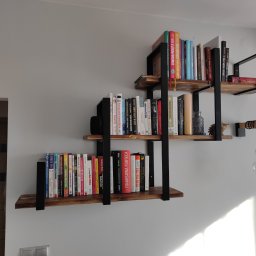 FilART - Construction Damian Filar - Trzy półki z drewna i metalu w stylu industrialnym zamontowane na szarej ścianie, wypełnione książkami o różnej tematyce i kolorowych okładkach, oświetlone światłem dziennym wpadającym z prawej...