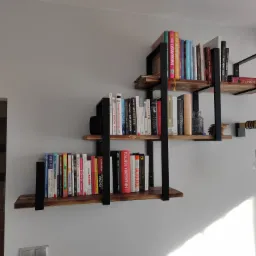 Trzy półki z drewna i metalu w stylu industrialnym zamontowane na szarej ścianie, wypełnione książkami o różnej tematyce i kolorowych okładkach, oświetlone światłem dziennym wpadającym z prawej...