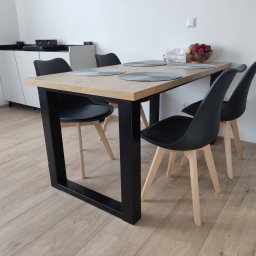 FilART - Construction Damian Filar - Stół z blatem z jasnego drewna i czarnymi, metalowymi nogami, otoczony czterema czarnymi krzesłami z jasnymi, drewnianymi nogami; na blacie okrągłe podkładki i misa z owocami.