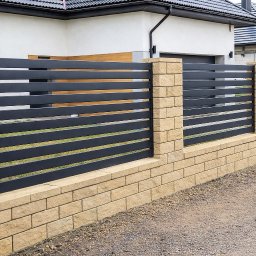 FilART - Construction Damian Filar - Nowoczesne ogrodzenie z poziomych, grafitowych paneli metalowych, osadzone na niskim murku z beżowych bloczków. Słupki z tego samego materiału. Dom w tle.