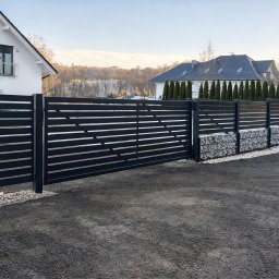 FilART - Construction Damian Filar - Nowoczesne, poziome ogrodzenie panelowe w kolorze antracytowym z gabionami u podstawy, na tle domu i zieleni. Widok z zewnątrz posesji.