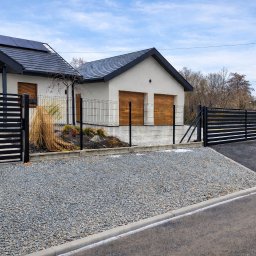 FilART - Construction Damian Filar - Nowoczesna posesja z czarnym ogrodzeniem panelowym i przesuwną bramą wjazdową. Widoczny fragment domu z panelami słonecznymi i żwirowy podjazd.