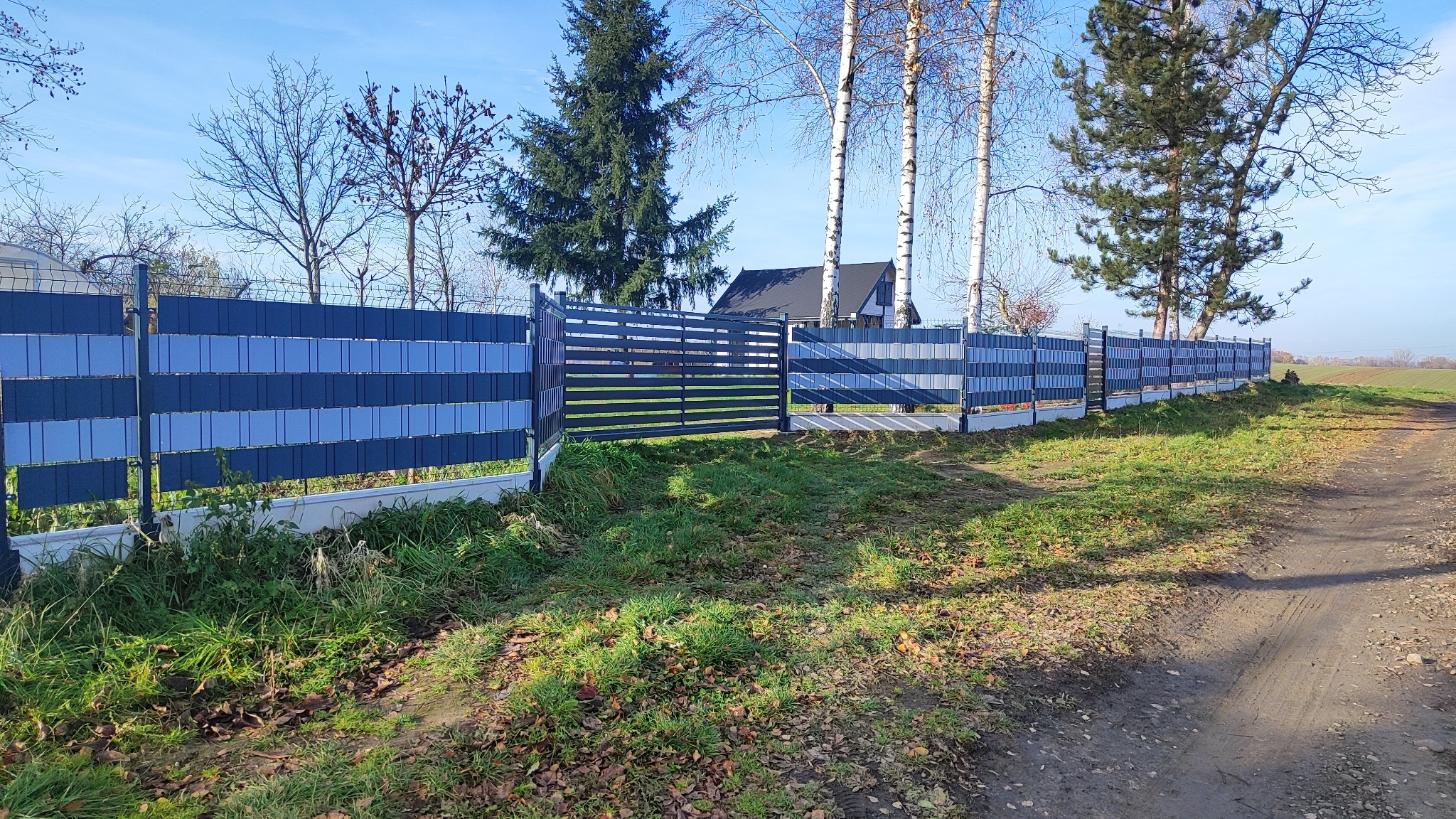 Nowoczesne ogrodzenie panelowe z poziomą bramą w kolorach szaro-niebieskich, widok z zewnątrz, z polną drogą i drzewami w tle.