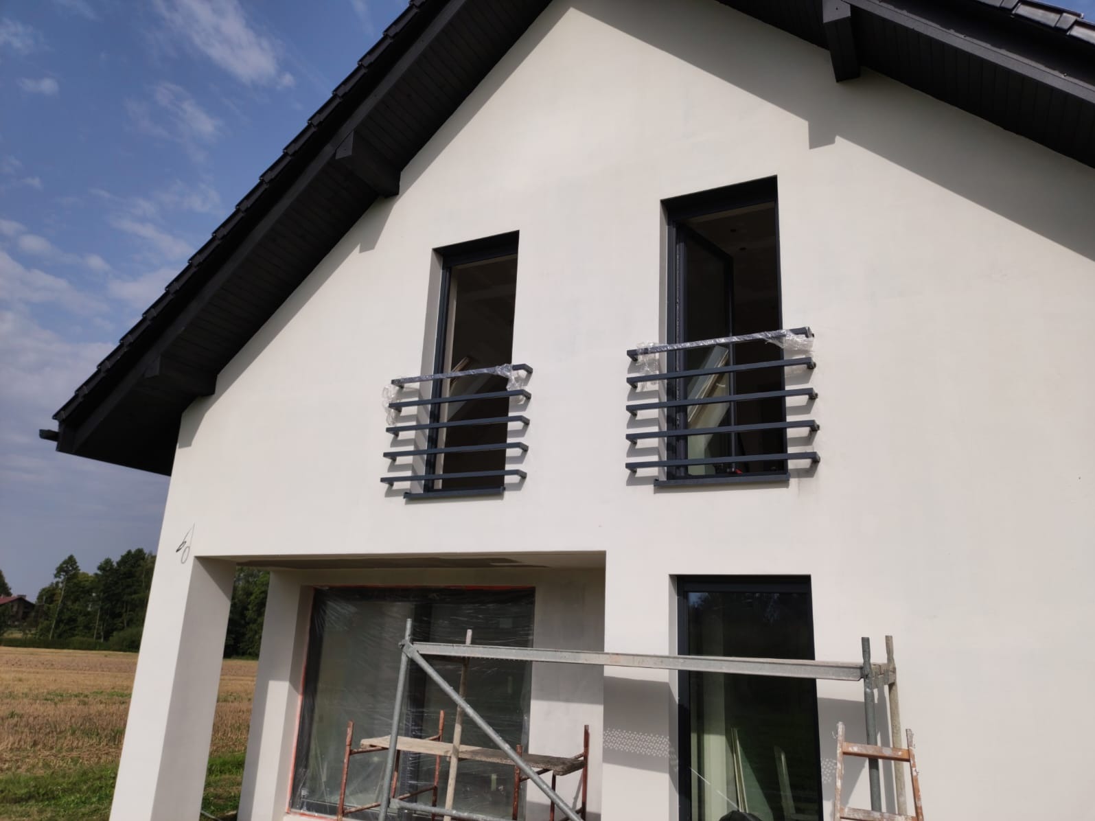 Nowoczesny dom jednorodzinny z białymi ścianami i czarną stolarką okienną, z zamontowanymi metalowymi balustradami okiennymi, częściowo jeszcze zabezpieczonymi folią ochronną, widoczne rusztowanie...