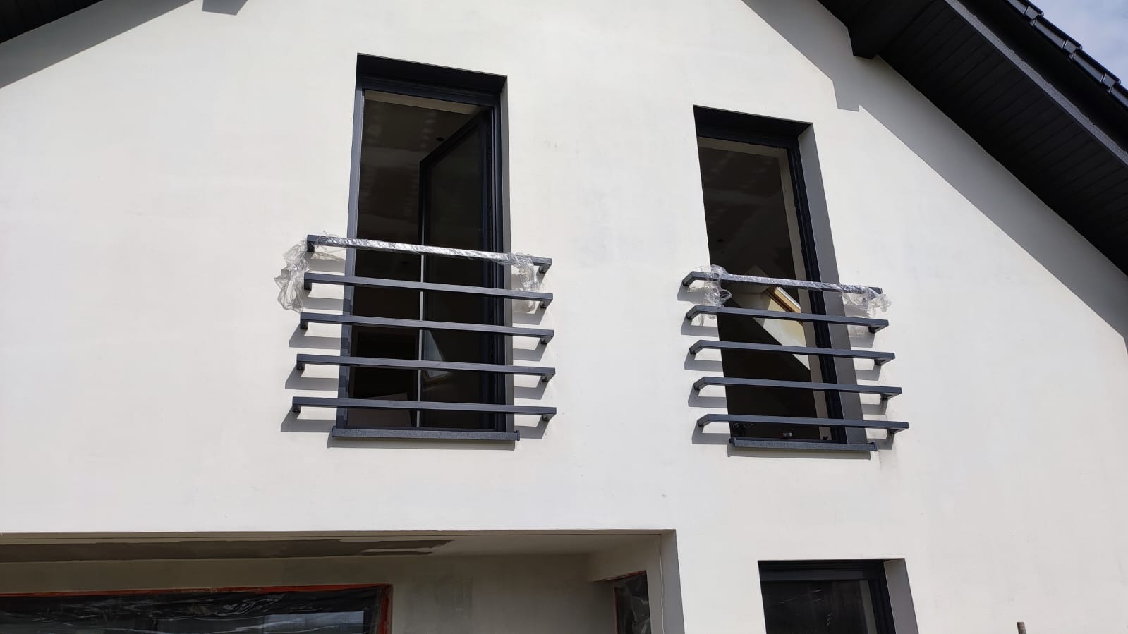 Nowoczesne, metalowe balustrady balkonowe z poziomymi prętami, częściowo zabezpieczone folią ochronną, zamontowane przy oknach na elewacji budynku w trakcie wykończenia.