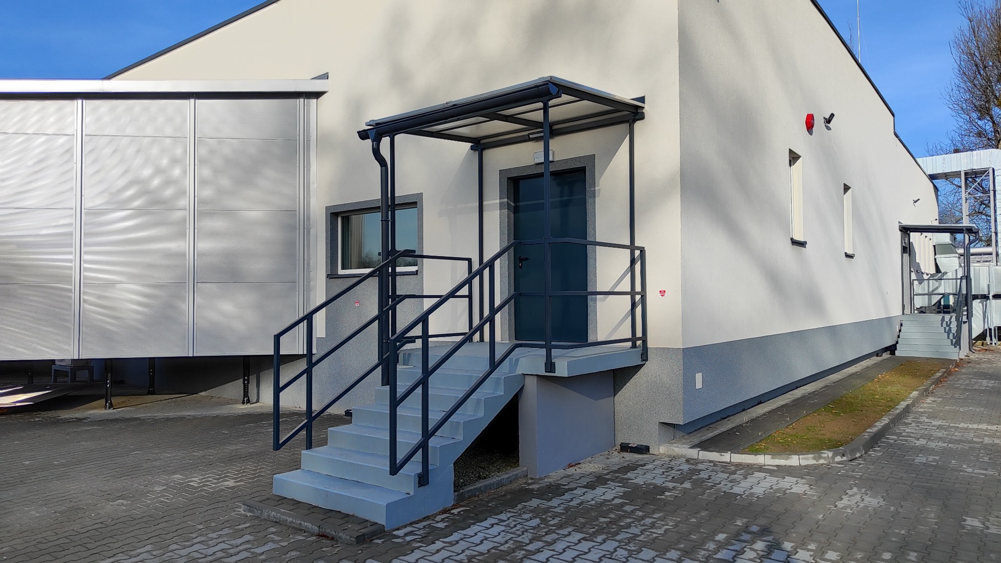 Szare schody z metalową balustradą prowadzące do ciemnozielonych drzwi wejściowych z daszkiem, widok z zewnątrz na tle jasnego budynku przemysłowego z blachy falistej.