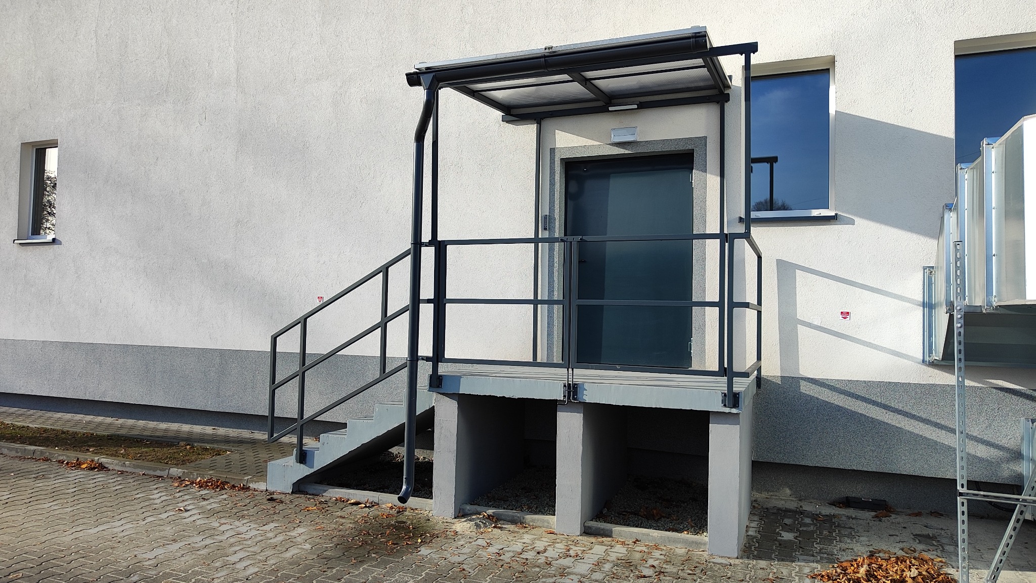 Zewnętrzne schody z czarną metalową balustradą prowadzące do drzwi zadaszonych metalową konstrukcją, obok okna i instalacja wentylacyjna na tle białej ściany budynku.