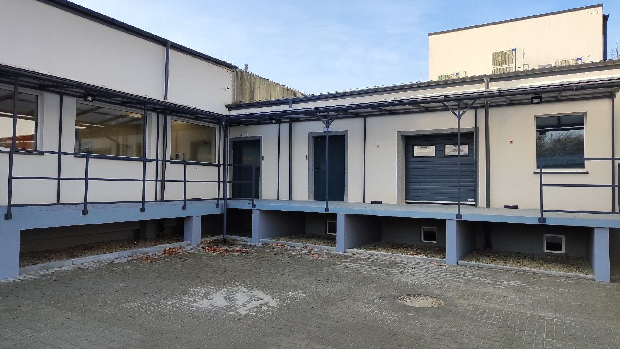 Rampa załadunkowa z trzema wejściami: dwoje drzwi i segmentowa brama garażowa w kolorze grafitowym, zadaszona konstrukcja metalowa, ściany w kolorze kremowym.