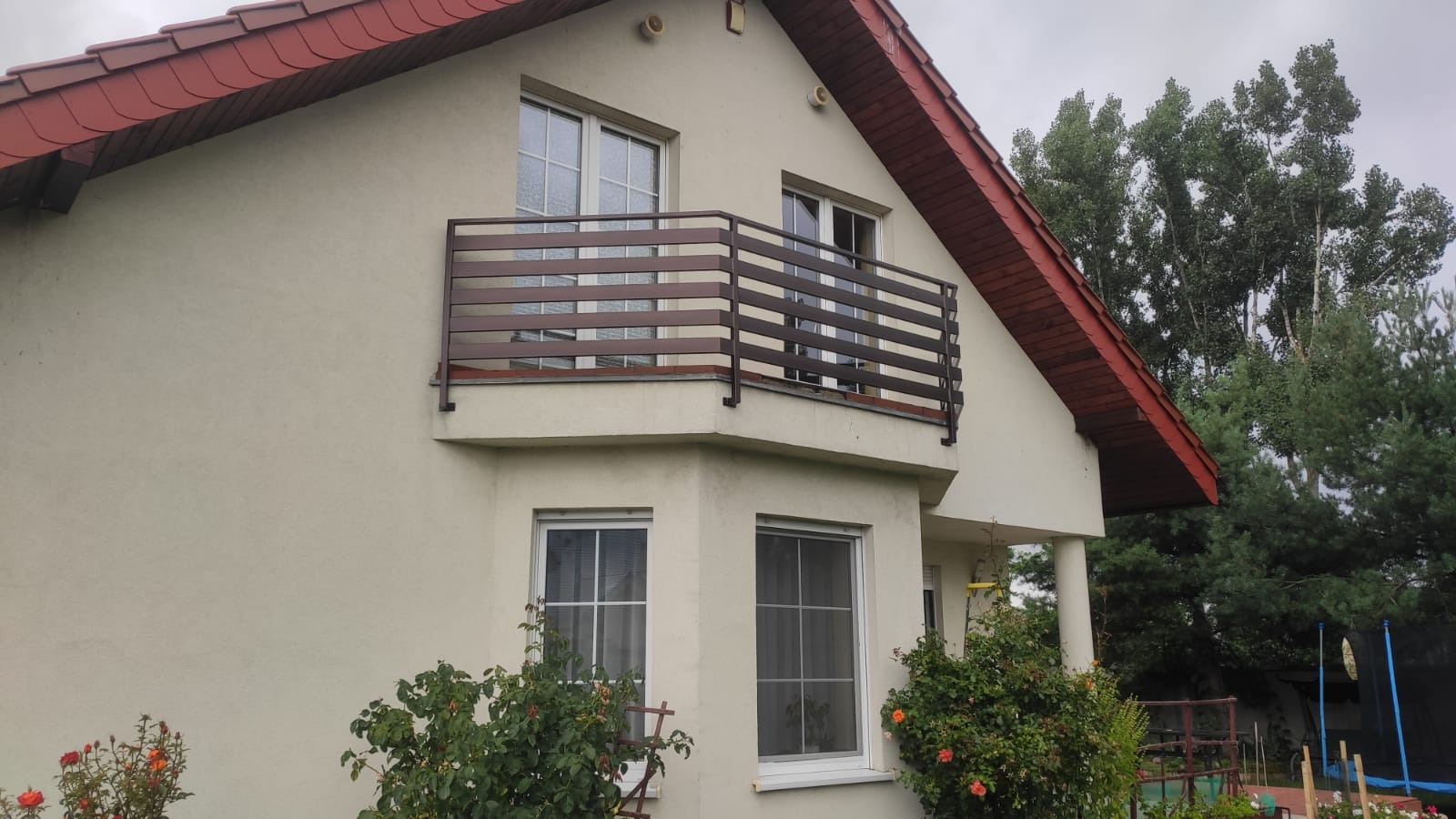 Dom jednorodzinny z beżową elewacją i balkonem z brązową, poziomą balustradą, widok z ogrodu z kwitnącymi różami.