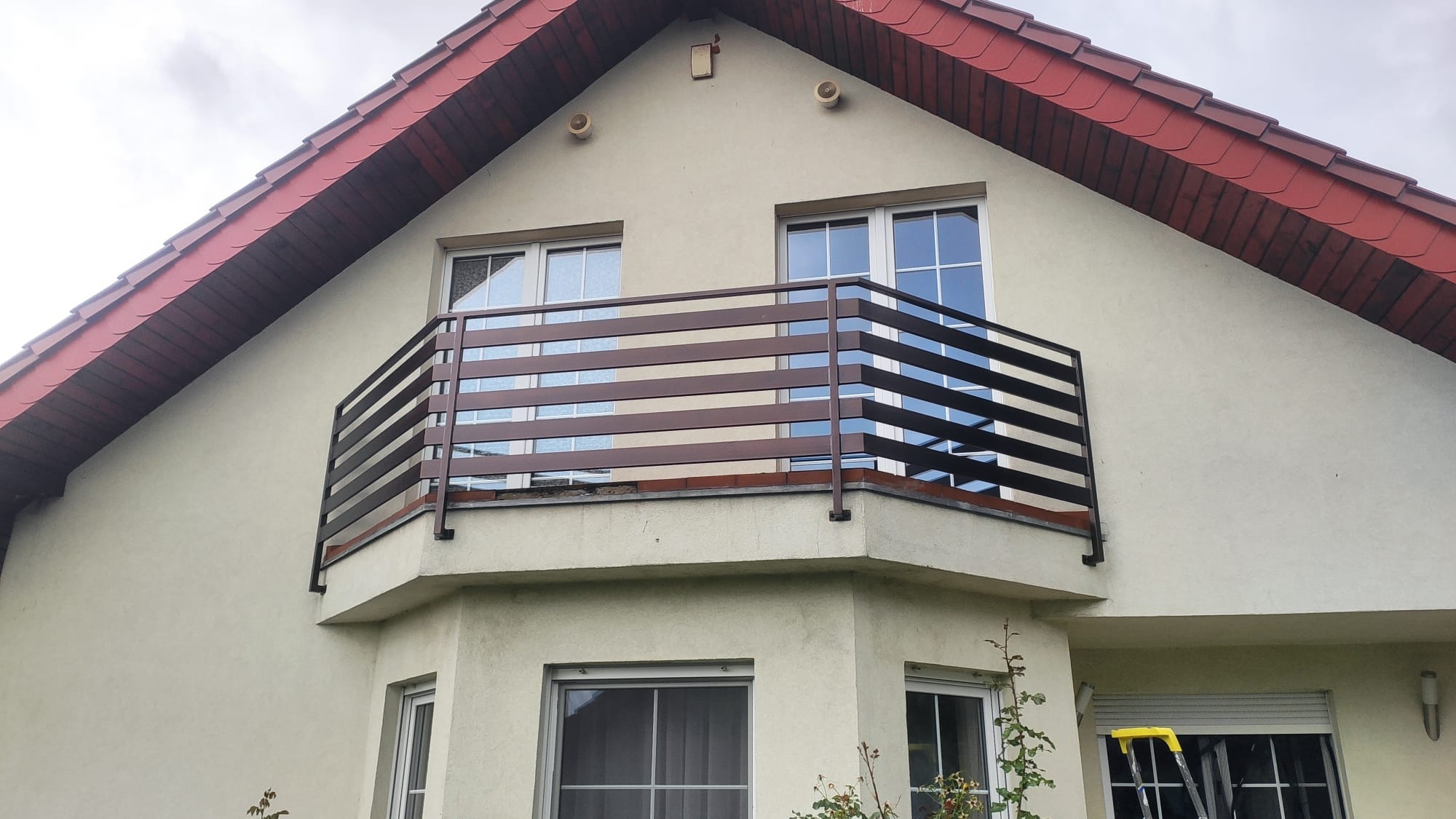 Balkon z nowoczesną, horyzontalną balustradą metalową w kolorze ciemnego brązu, zamontowaną w elewacji domu z czerwoną dachówką, widok z dołu na tle pochmurnego nieba.