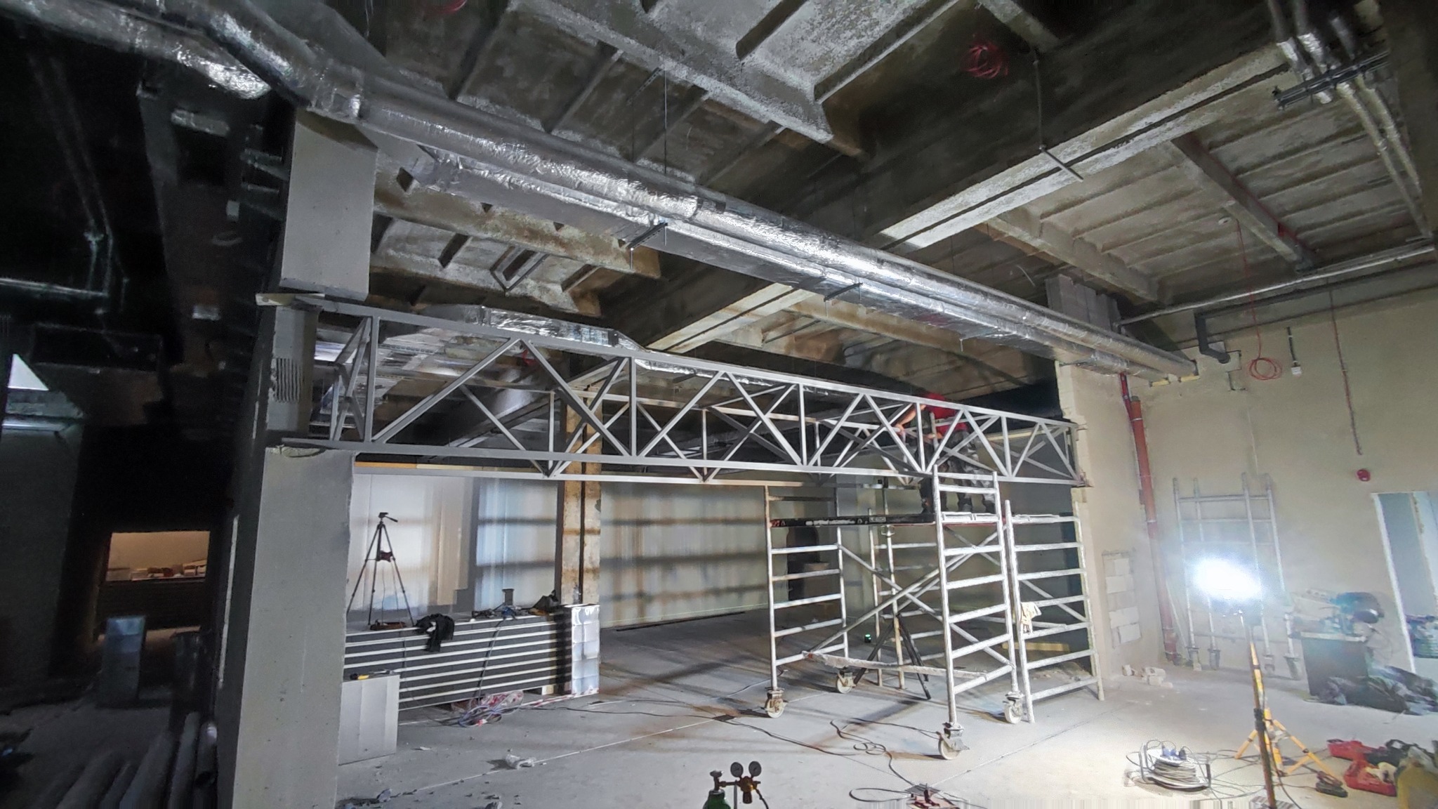 Wnętrze hali przemysłowej w trakcie budowy, widoczna stalowa konstrukcja kratowa pod sufitem, rusztowania na kółkach, instalacje wentylacyjne i oświetlenie budowlane.