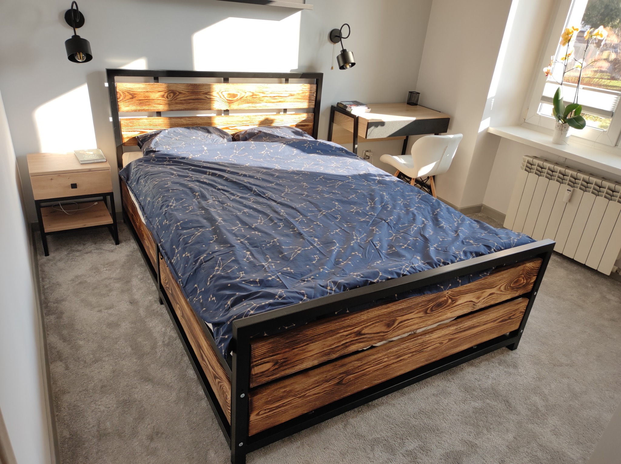 Drewniane łóżko z metalową ramą i pościelą w gwiazdy, widok z dołu, w tle biurko i okno z kwiatami.