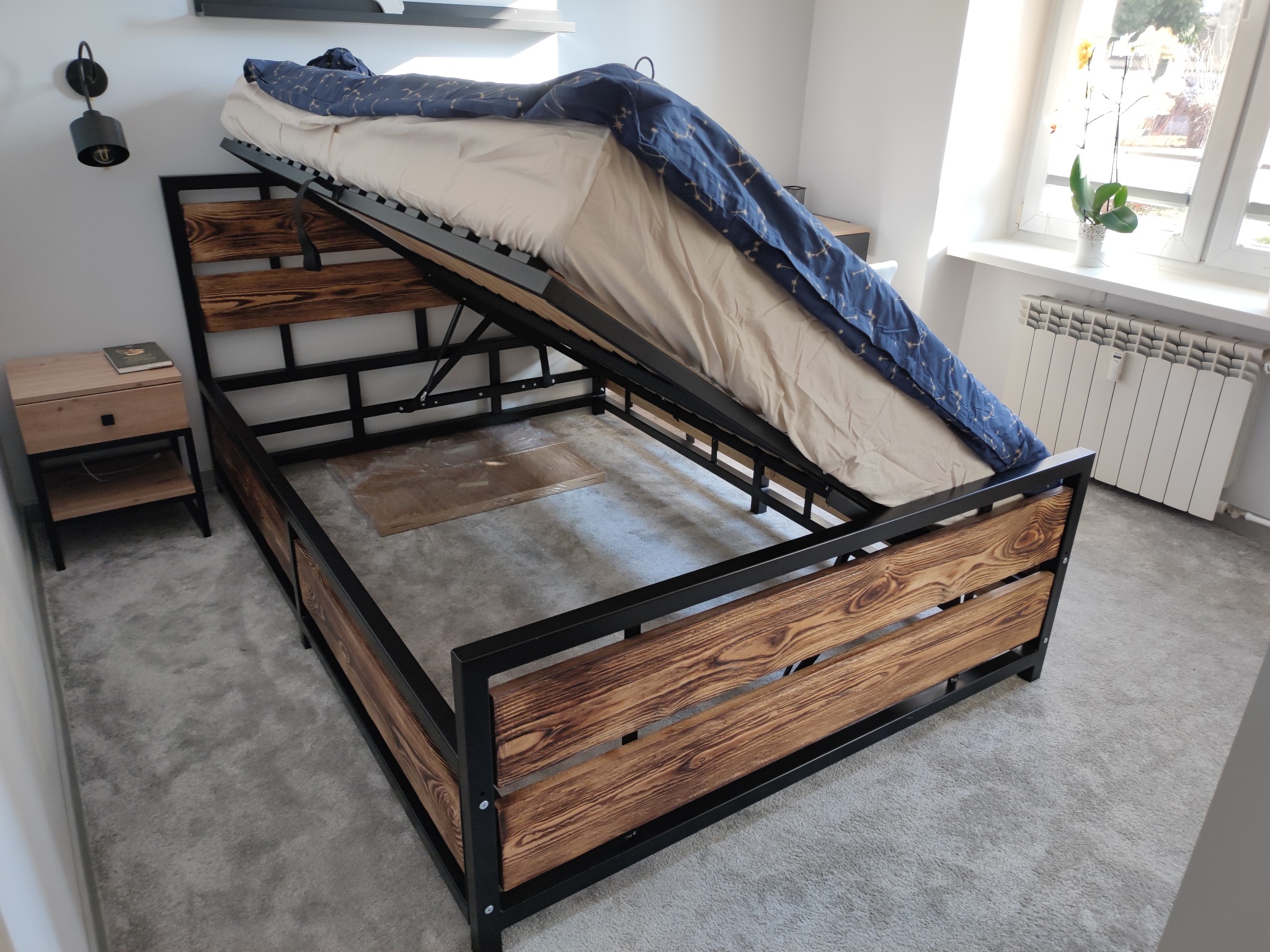 Loftowe łóżko z podnoszonym stelażem, z widoczną drewnianą ramą z palonego drewna i czarną metalową konstrukcją, częściowo podniesione, ukazujące przestrzeń do przechowywania, z pościelą...