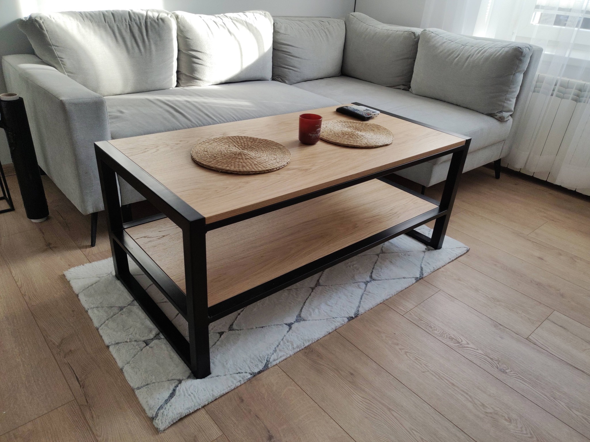 Stylowy stolik kawowy z drewnianym blatem i czarną metalową ramą, na puszystym dywanie w salonie, obok szarej sofy. Widoczne dekoracje na blacie.