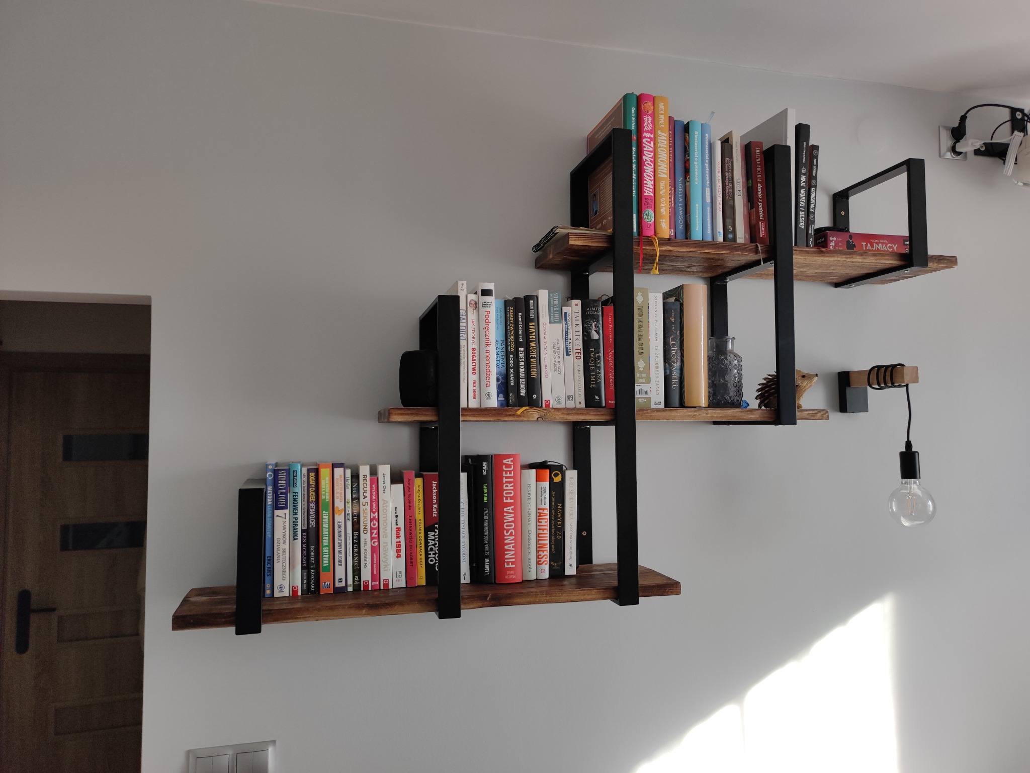 Trzy półki z drewna i metalu w stylu industrialnym zamontowane na szarej ścianie, wypełnione książkami o różnej tematyce i kolorowych okładkach, oświetlone światłem dziennym wpadającym z prawej...