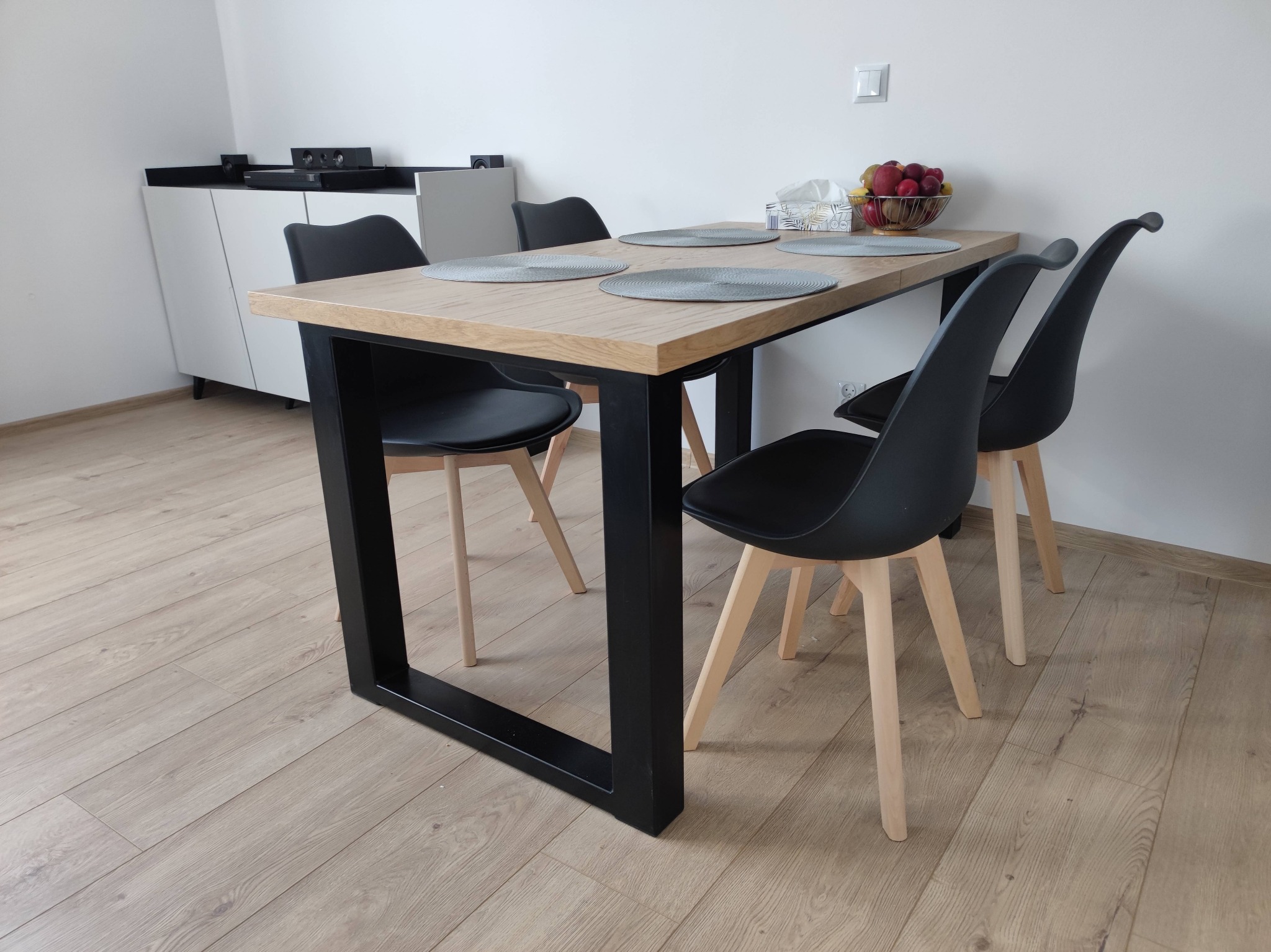 Stół z blatem z jasnego drewna i czarnymi, metalowymi nogami, otoczony czterema czarnymi krzesłami z jasnymi, drewnianymi nogami; na blacie okrągłe podkładki i misa z owocami.