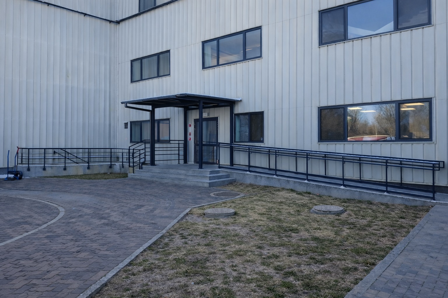 Metalowa balustrada przy wejściu do budynku z rampą dla niepełnosprawnych. Ciemna konstrukcja na tle jasnej elewacji. Widoczne okna i chodnik z kostki.