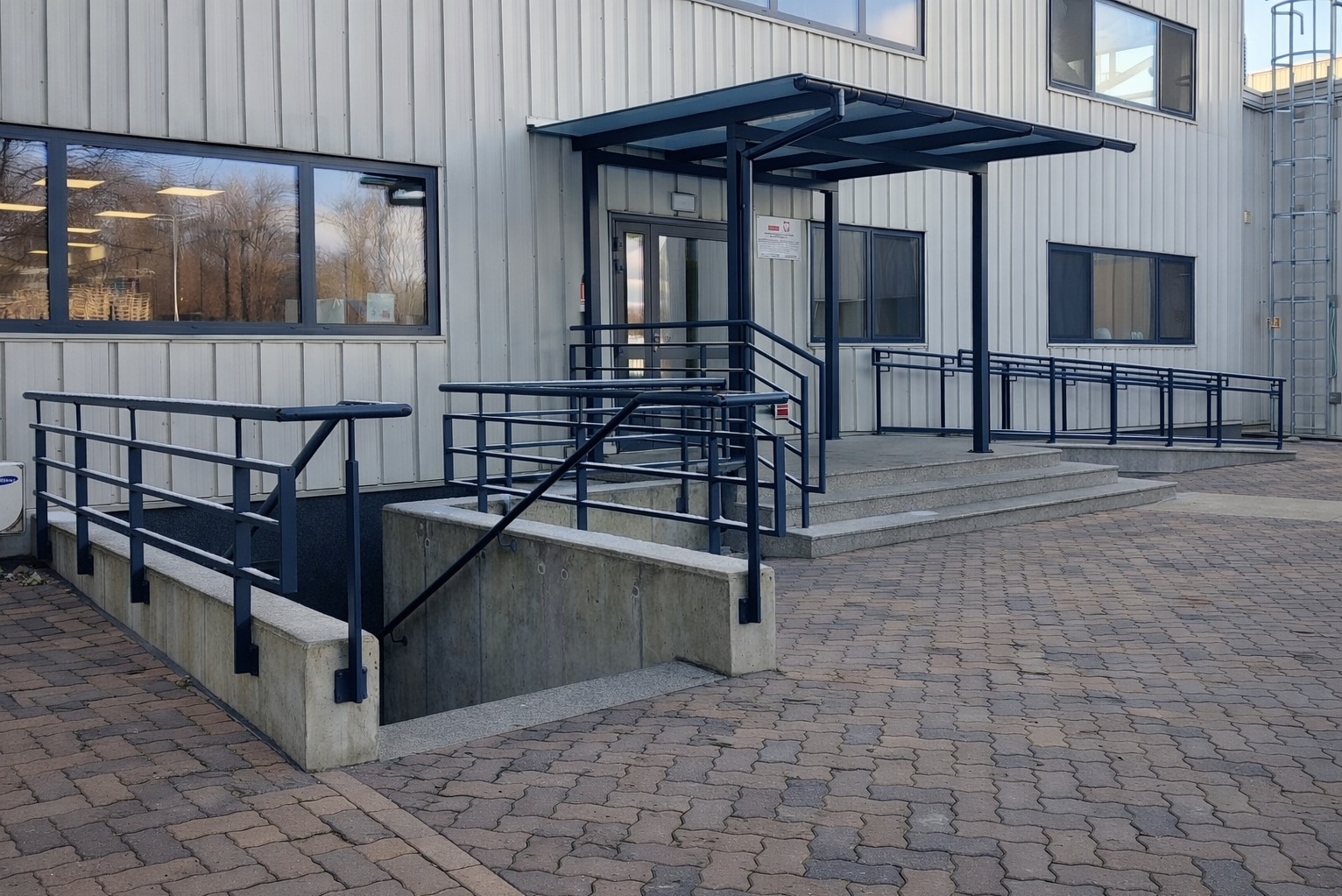Zewnętrzne schody betonowe z metalową balustradą prowadzące do budynku o metalowej elewacji. Widoczny podjazd dla wózków inwalidzkich i zadaszenie nad wejściem.