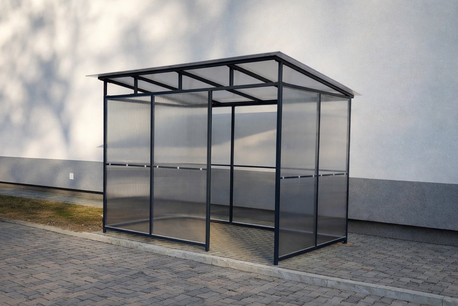 Nowoczesna, metalowa wiata z przezroczystymi ścianami, stojąca na kostce brukowej przy jasnej ścianie budynku. Minimalistyczny design, widoczne detale konstrukcyjne.