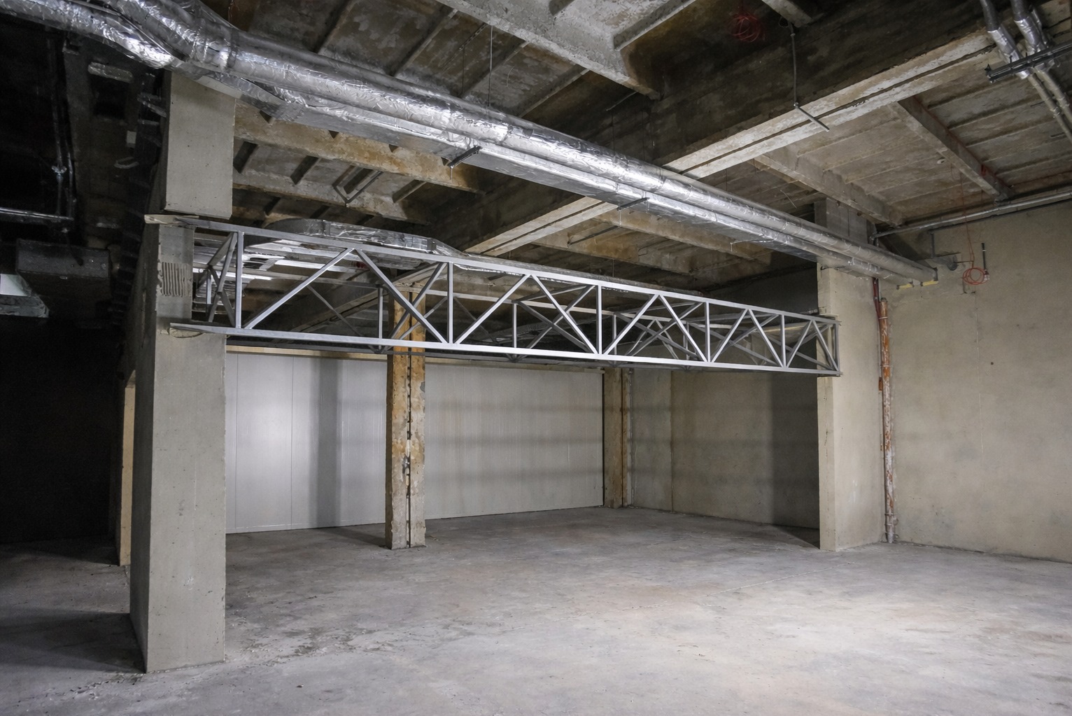 Surowe wnętrze z betonowymi filarami i metalową konstrukcją pod sufitem. Widoczne rury wentylacyjne i surowy beton. Minimalistyczny, industrialny styl.