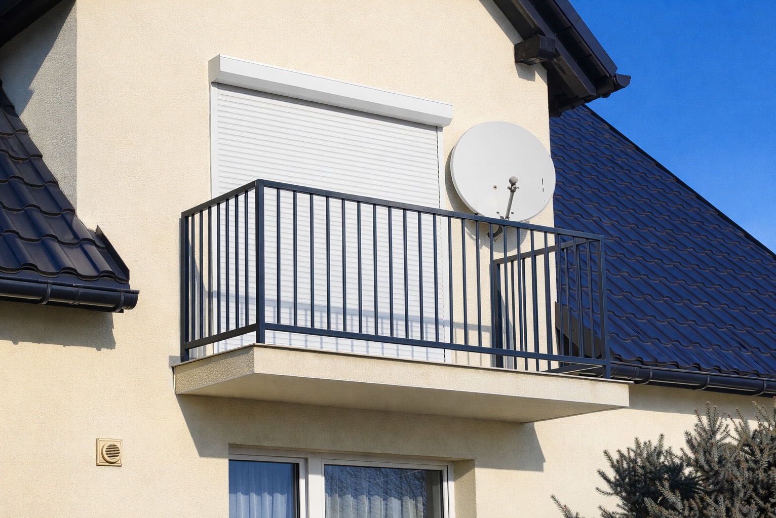 Nowoczesna, czarna balustrada balkonowa z pionowymi szczebelkami, zamontowana na balkonie domu z beżową elewacją i białą roletą okienną. Widoczna antena satelitarna.
