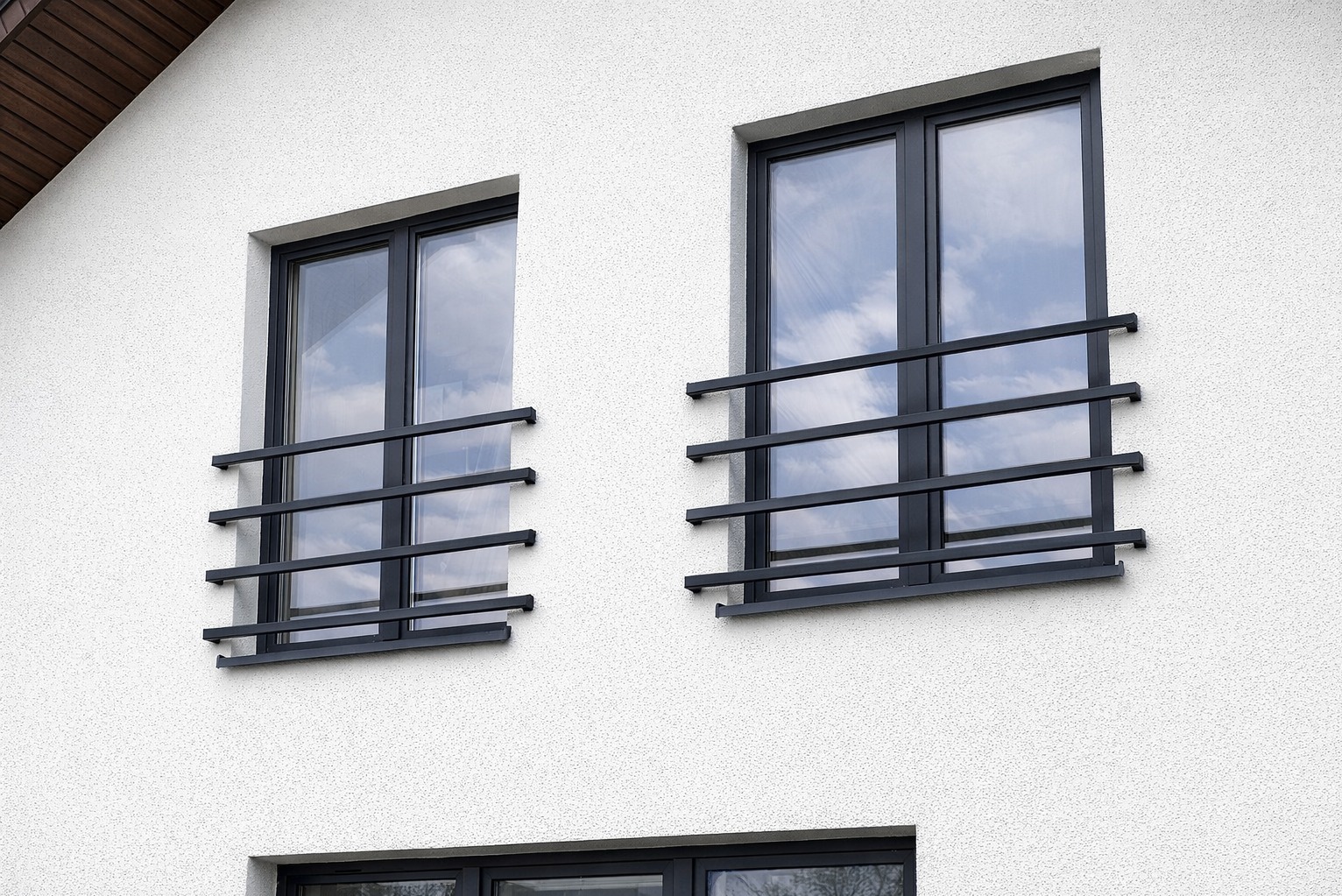 Dwa okna z czarnymi, minimalistycznymi kratami na białej, teksturowanej elewacji budynku. Widoczne odbicie nieba w szybach.
