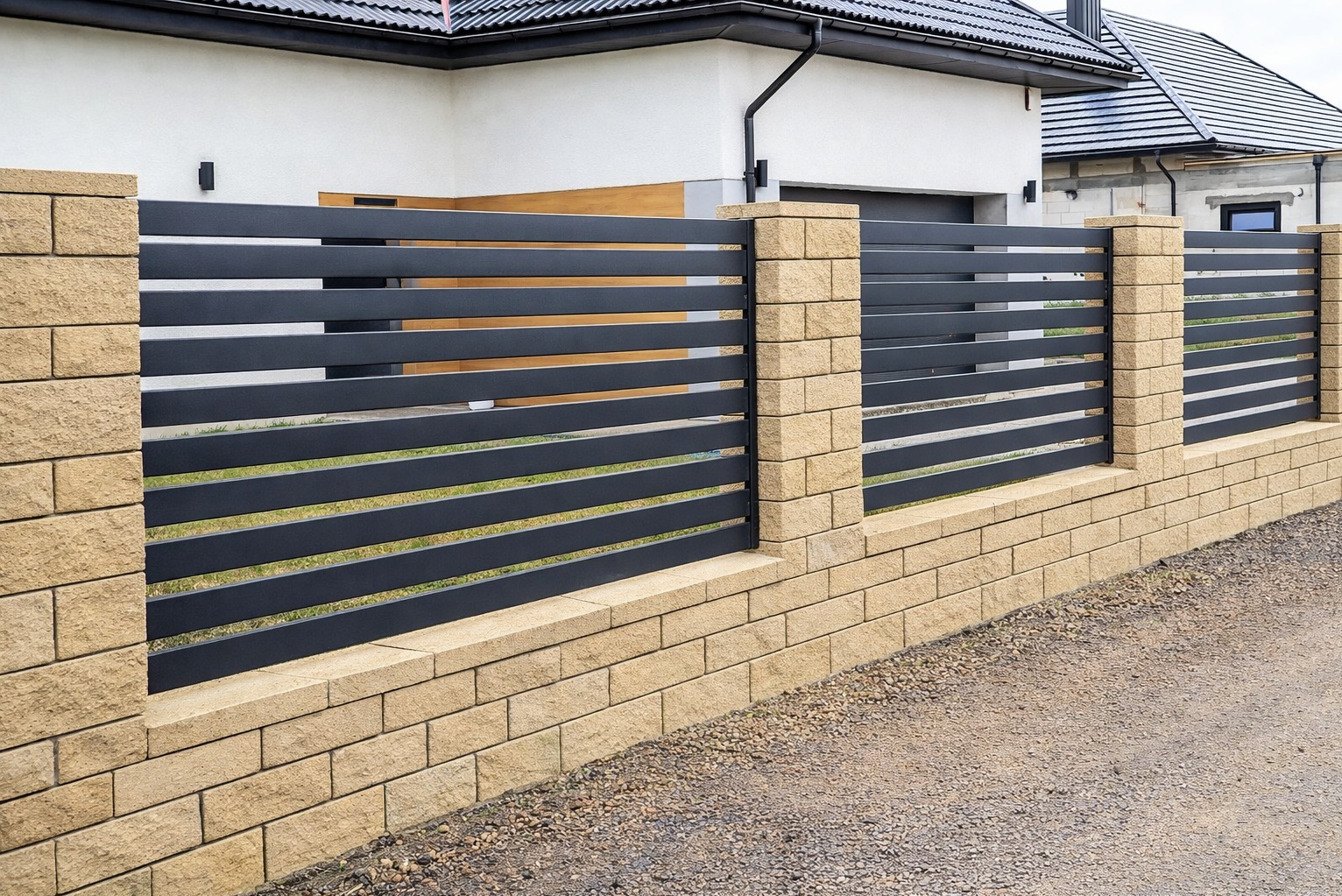 Nowoczesne ogrodzenie z poziomych, grafitowych paneli metalowych, osadzone na niskim murku z beżowych bloczków. Słupki z tego samego materiału. Dom w tle.