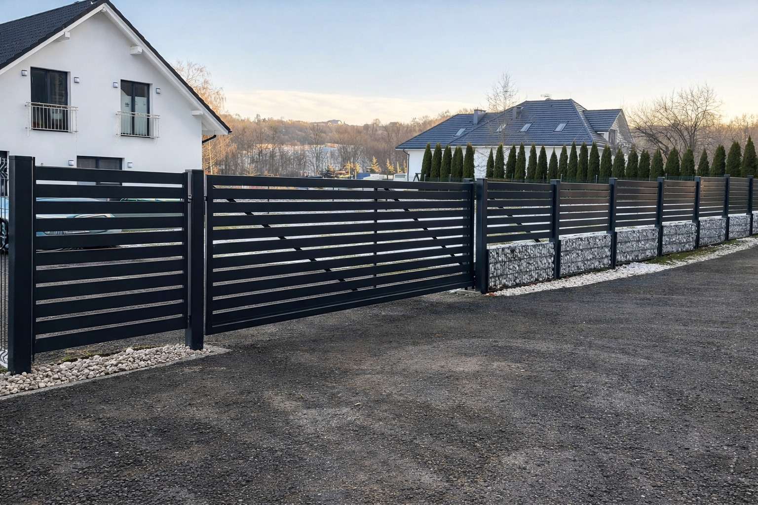 Nowoczesne, poziome ogrodzenie panelowe w kolorze antracytowym z gabionami u podstawy, na tle domu i zieleni. Widok z zewnątrz posesji.