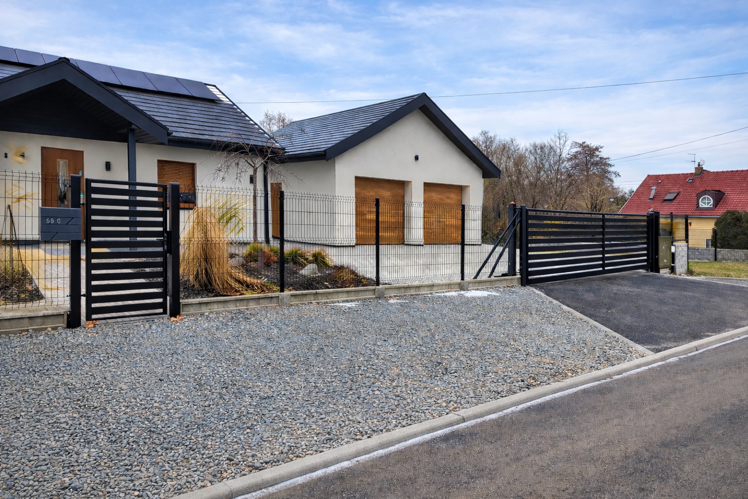 Nowoczesna posesja z czarnym ogrodzeniem panelowym i przesuwną bramą wjazdową. Widoczny fragment domu z panelami słonecznymi i żwirowy podjazd.