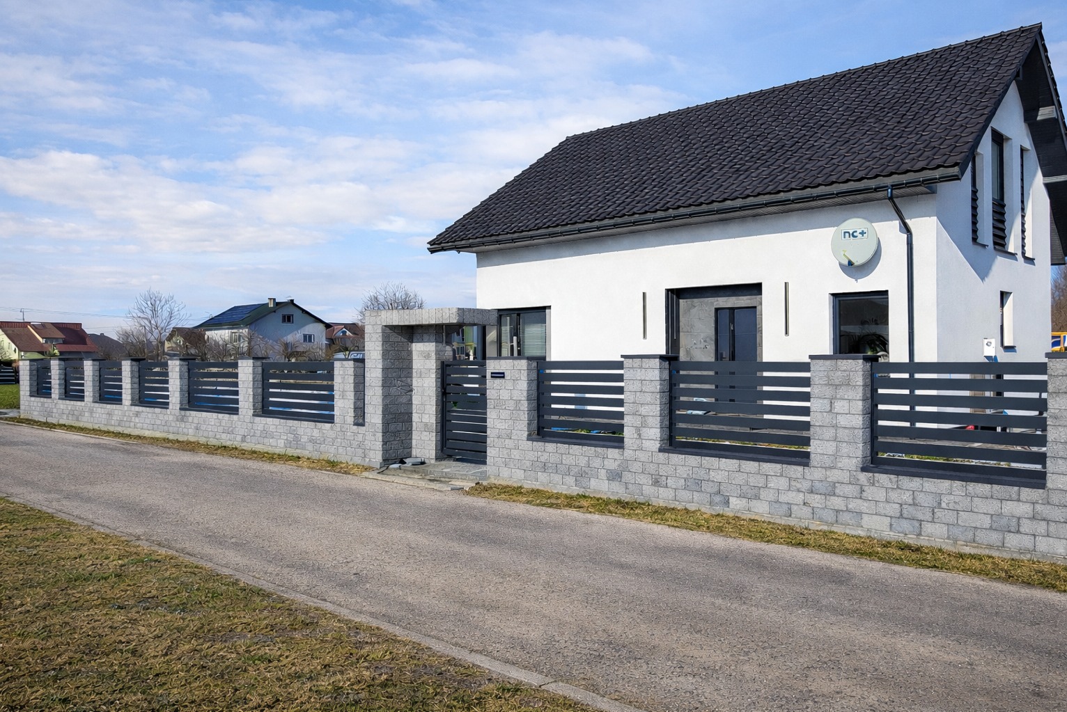 Nowoczesne ogrodzenie panelowe z szarej kostki i poziomych, grafitowych przęseł, otaczające posesję z białym domem. Brama wjazdowa wkomponowana w ogrodzenie.