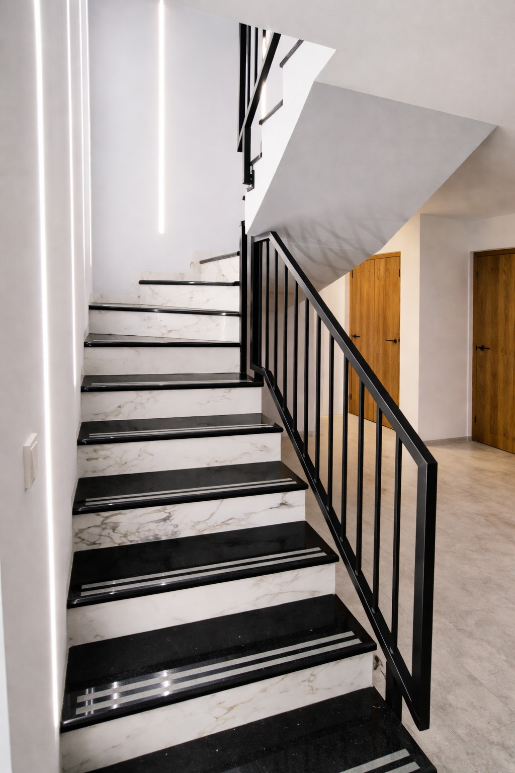 Nowoczesne schody z czarno-białymi stopniami marmurowymi i czarną metalową balustradą. Oświetlenie LED wzdłuż ściany. Widoczne dwie drewniane drzwi w tle.