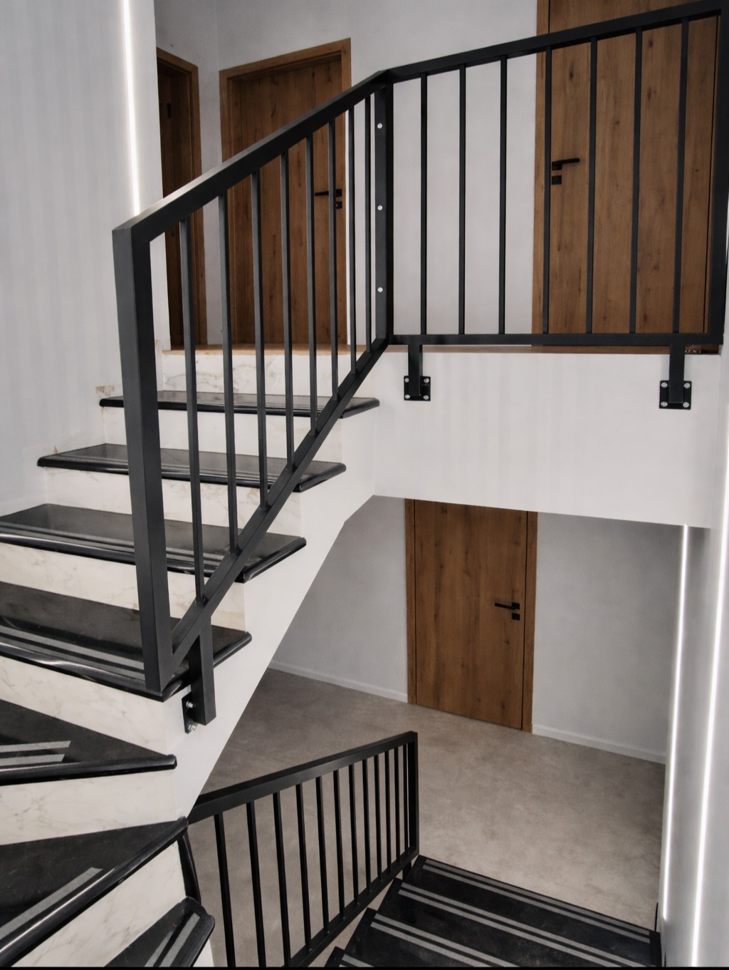 Nowoczesne schody z czarną metalową balustradą i marmurowymi stopniami. Widoczne trzy drewniane drzwi w tle. Minimalistyczny design, oświetlenie LED.