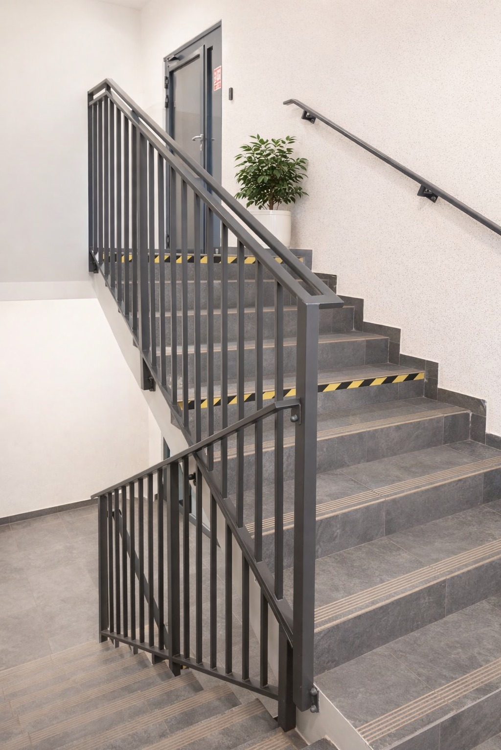 Klatka schodowa z szarymi, metalowymi balustradami i płytkami. Na schodach żółto-czarne pasy antypoślizgowe. Na podeście doniczka z zieloną rośliną. Minimalistyczny design.