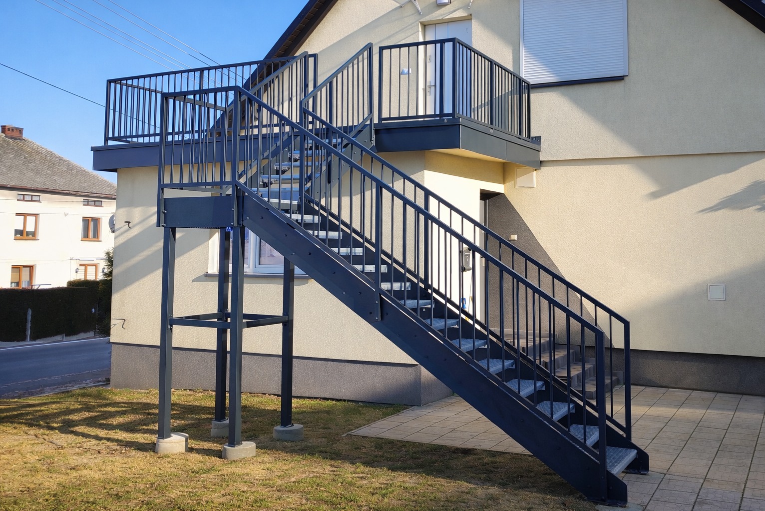 Zewnętrzne schody metalowe z balustradą prowadzące na balkon, pomalowane na ciemny kolor, zamontowane przy jasnym budynku. Widok z dołu, w słoneczny dzień.