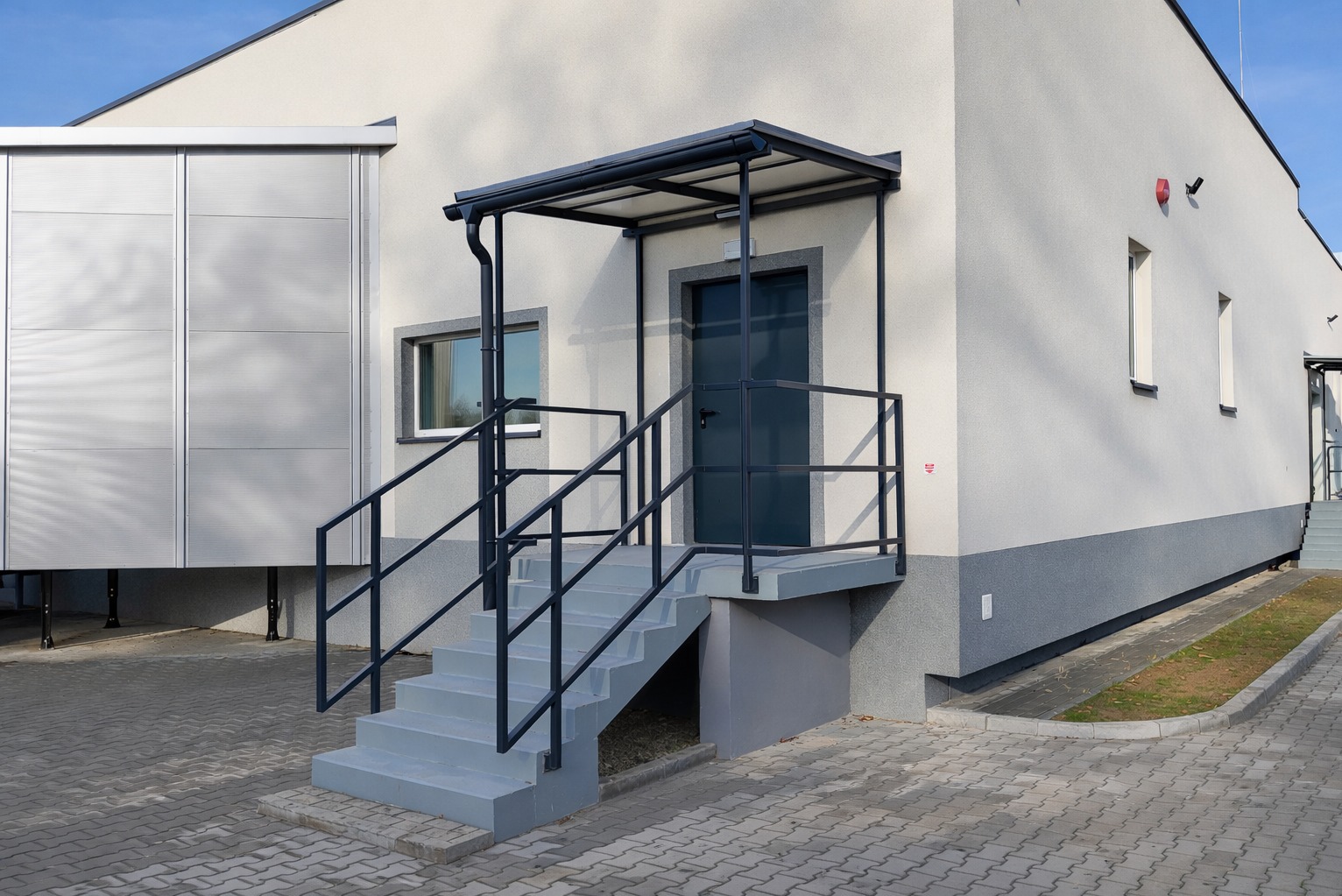 Zewnętrzne schody metalowe z balustradą i zadaszeniem prowadzące do drzwi wejściowych budynku. Szare stopnie i ciemna stalowa konstrukcja.