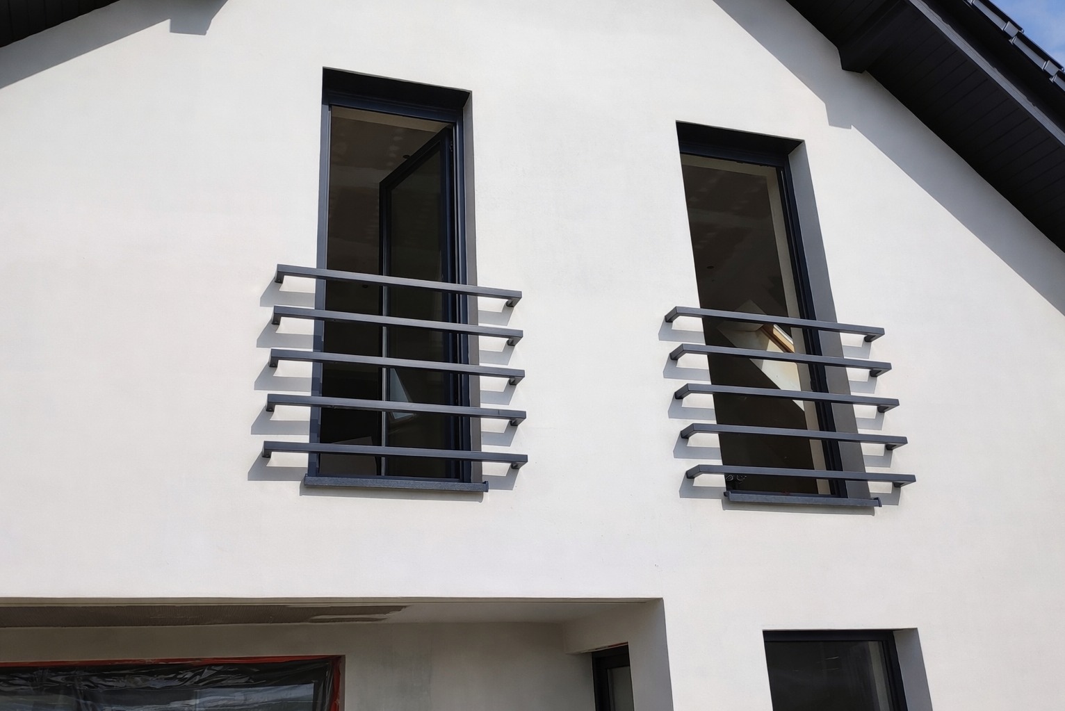 Nowoczesna, minimalistyczna balustrada okienna z ciemnego metalu na białej fasadzie domu z otwartym oknem. Geometryczny design, wyraźne cienie, surowy styl.