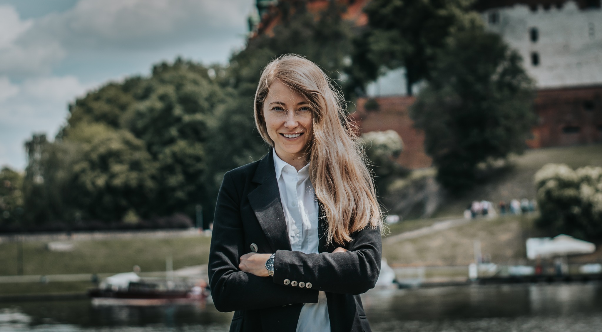 Uśmiechnięta kobieta w czarnej marynarce i białej koszuli, z zegarkiem na ręku, stoi na tle rzeki i zamku na wzgórzu.