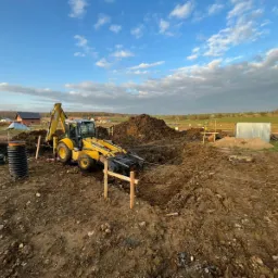 Żółta koparko-ładowarka w trakcie prac ziemnych na placu budowy, widoczne drewniane paliki wyznaczające obszar, w tle domy i pochmurne niebo.