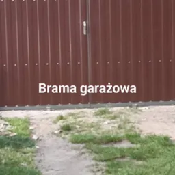Ciemnobrązowa brama wjazdowa z pionowych paneli metalowych, mocowana na nity, z widocznym fragmentem ceglanego muru w tle i ścieżką prowadzącą do bramy.