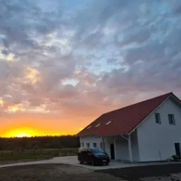 Nowo wybudowany dom jednorodzinny z czerwonym dachem i białą elewacją, sfotografowany o zachodzie słońca, z zaparkowanym czarnym vanem przed wejściem.