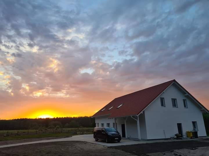 Nowo wybudowany dom jednorodzinny z czerwonym dachem i białą elewacją, sfotografowany o zachodzie słońca, z zaparkowanym czarnym vanem przed wejściem.