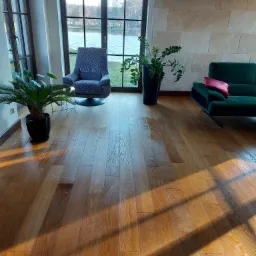 Jasne, lakierowane deski podłogowe w salonie z widokiem na jezioro, oświetlone popołudniowym słońcem tworzącym długie cienie, z elementami wystroju: zielona sofa z czerwoną poduszką, fotel...